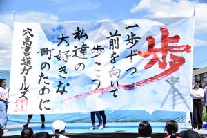 ステージで大きな紙に書いた書道作品を掲げる書道ガールズ。紙には「歩。一歩ずつ前を向いて歩こう。私たちの大好きな町のために」と書かれている。